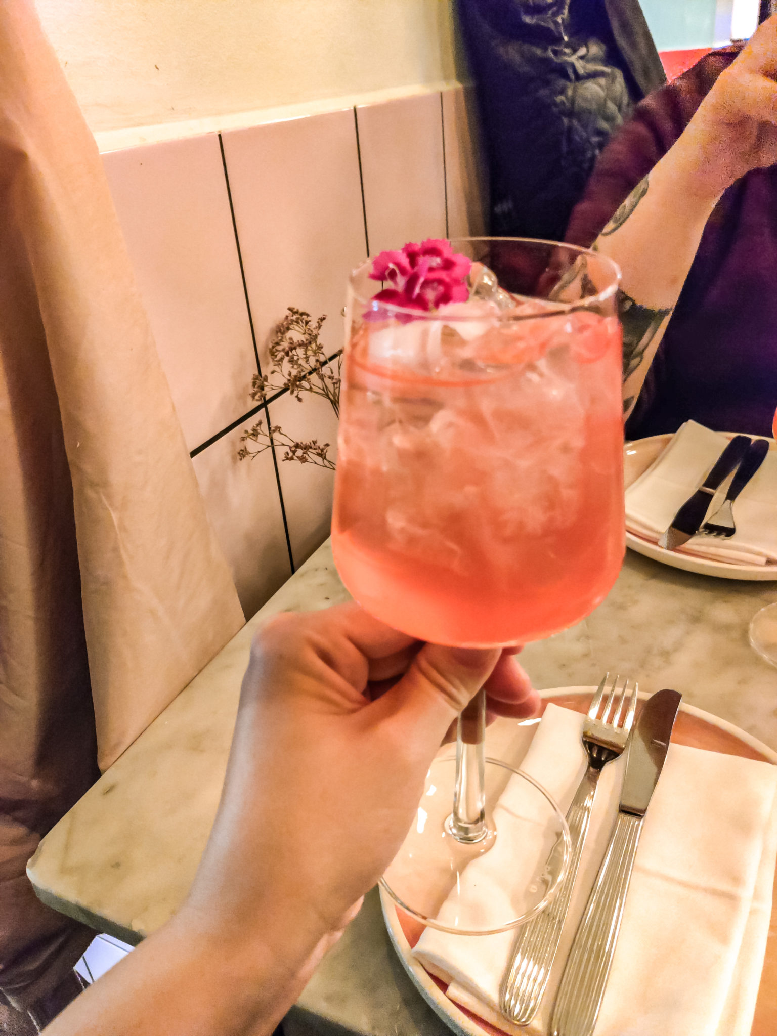 Käsi pitelee korkeajalkaisessa lasissa olevaa punertavaa, kukalla koristeltua juomaa.