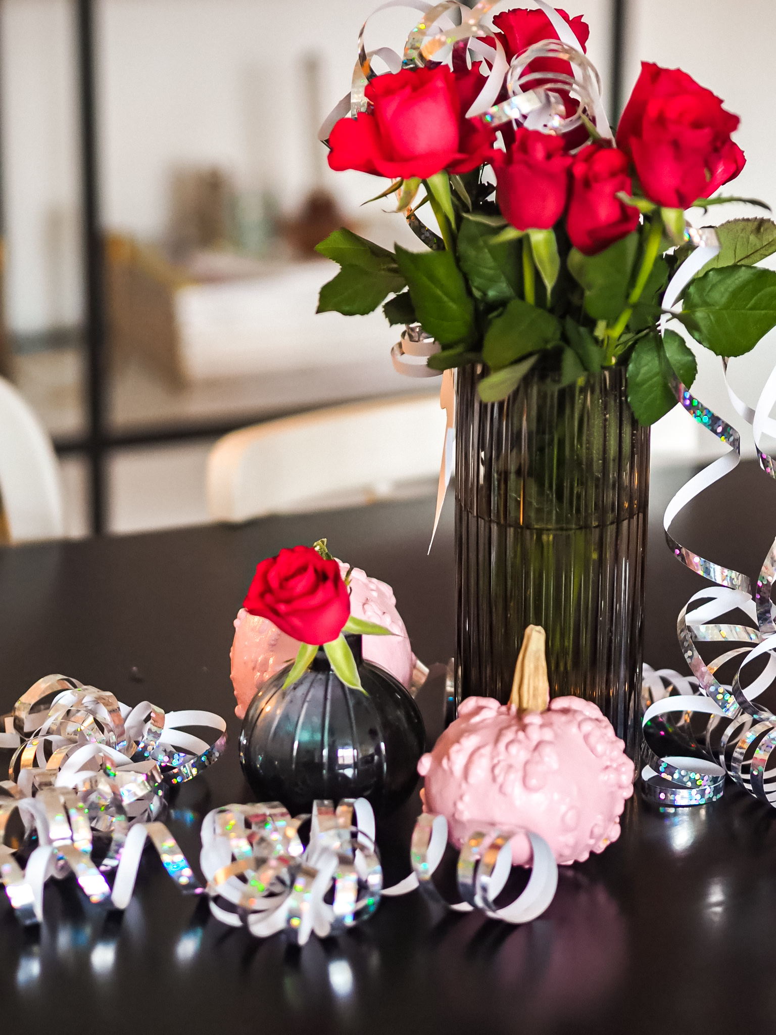 Maljakossa punaisia ruusuja ja pöydällä vaaleanpunaiseksi maalattuja kurpitsoja, sekä hopeista serpentiiniä.