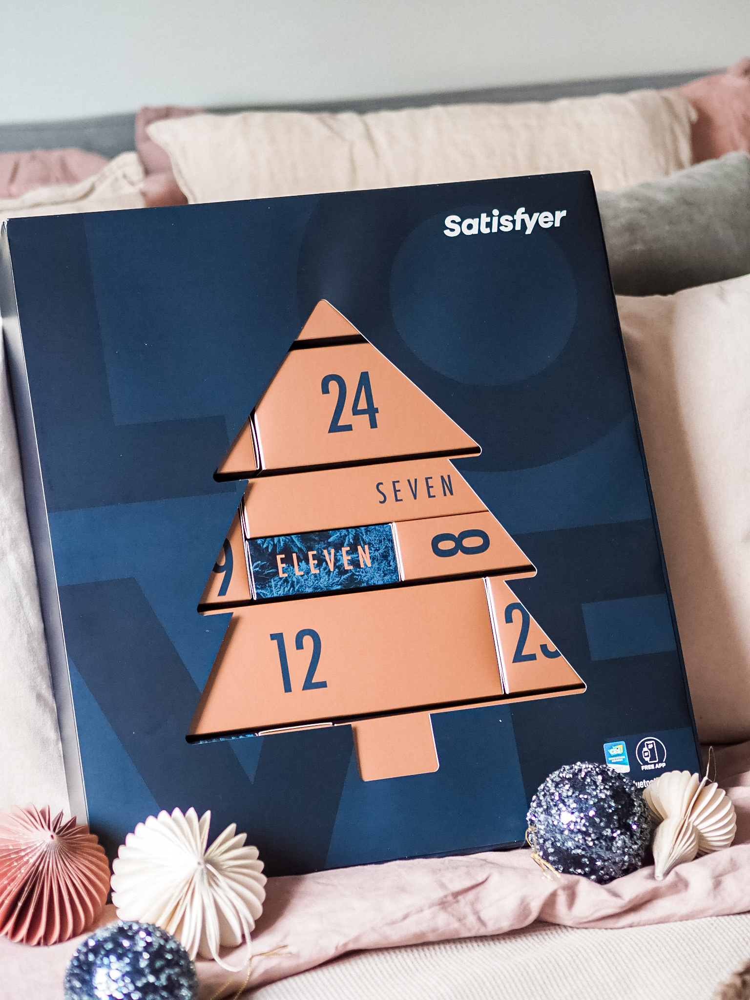 Satisfyer kalenteri on tummansininen. Päälli pahvissa on kuusen muotoinen aukko, josta näkee osan pronssin värisistä luukuista. Ympärillä koristeena joulupalloja.